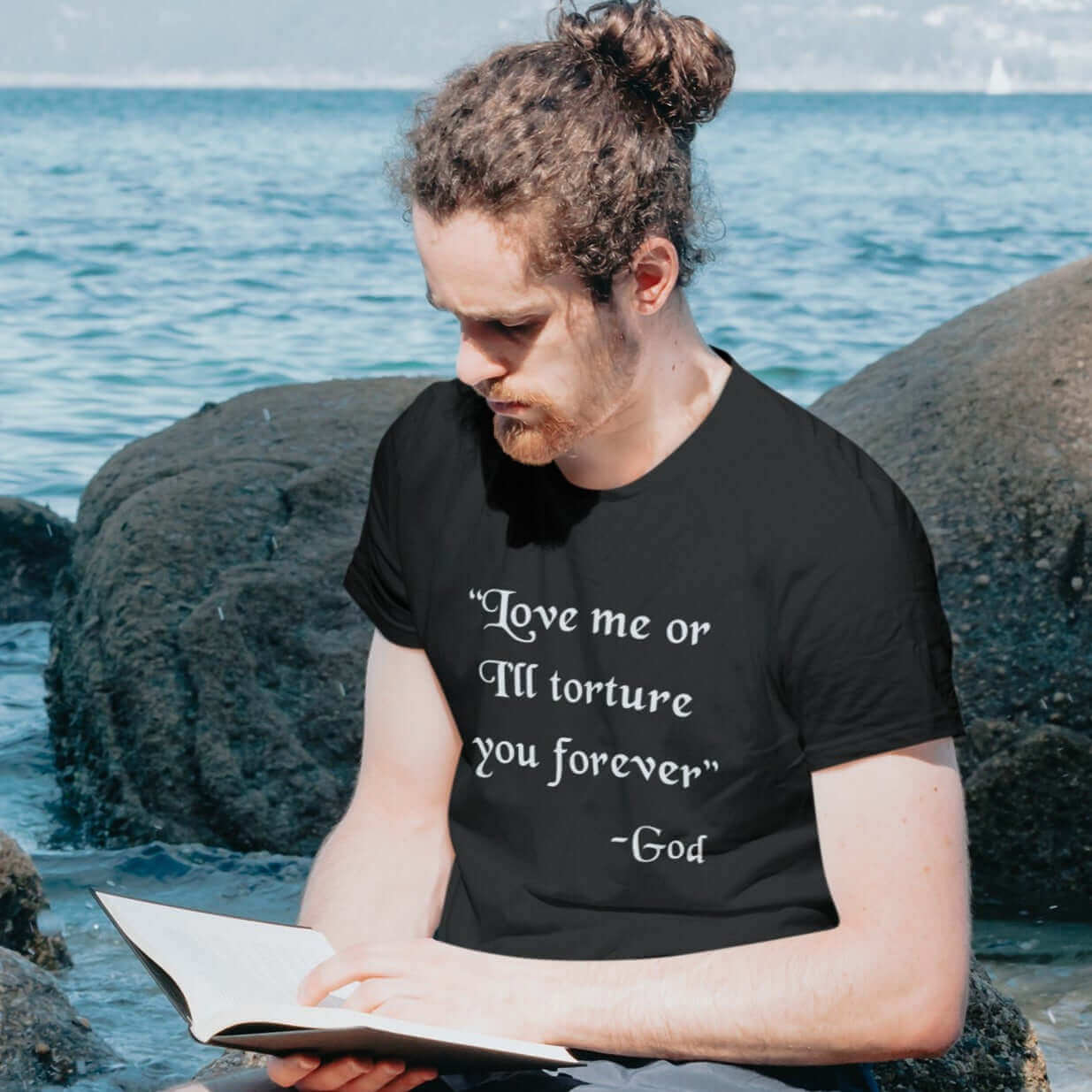 Man reading a book while wearing a black t-shirt with the quote Love me or I'll torture you forever-God printed on the front.