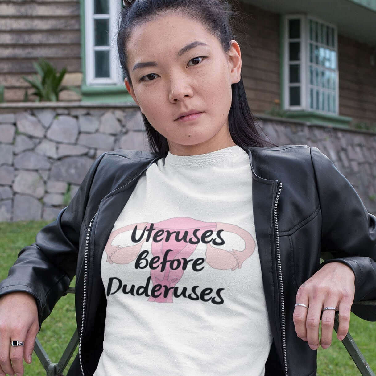 Woman leaning against a fence wearing a white t-shirt with image of a uterus and the words Uteruses before duderuses printed on the front.