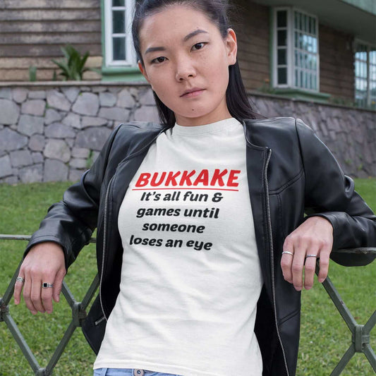 Serious looking woman leaning against a fence wearing white t-shirt with the words Bukkake it's all fun & games until someone loses an eye printed on the front.