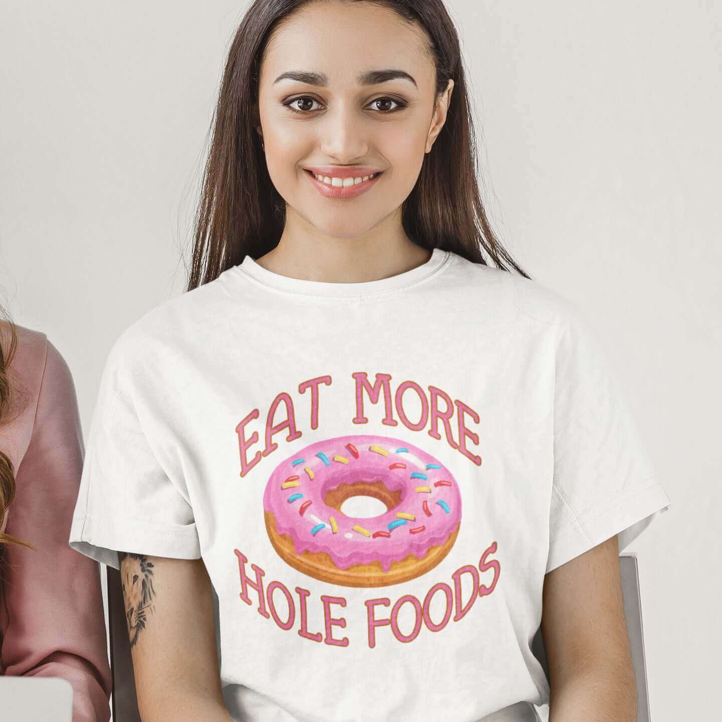 Woman wearing white t-shirt that has an image of a donut with pink icing and sprinkles and Eat more hole foods printed on the front.