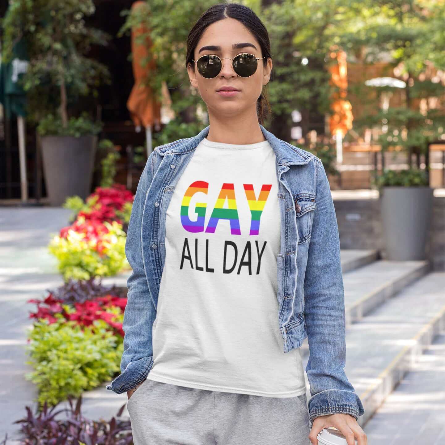 Woman wearing a white t-shirt with the words Gay all day printed on the front. The word Gay is in rainbow stripe font. 