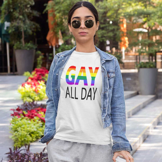 Woman wearing a white t-shirt with the words Gay all day printed on the front. The word Gay is in rainbow stripe font. 