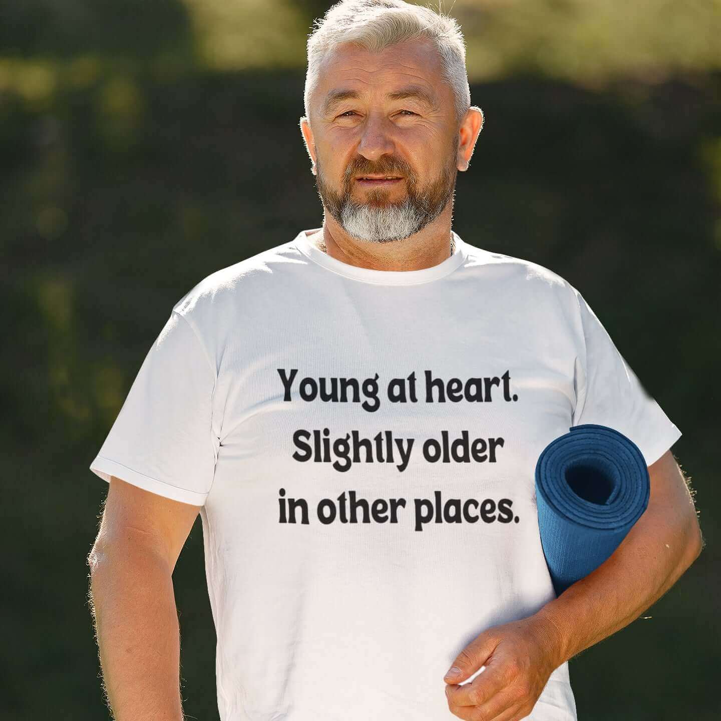 Older man carrying yoga mat wearing a white t-shirt with the words Young at heart, slightly older in other places printed on the front.