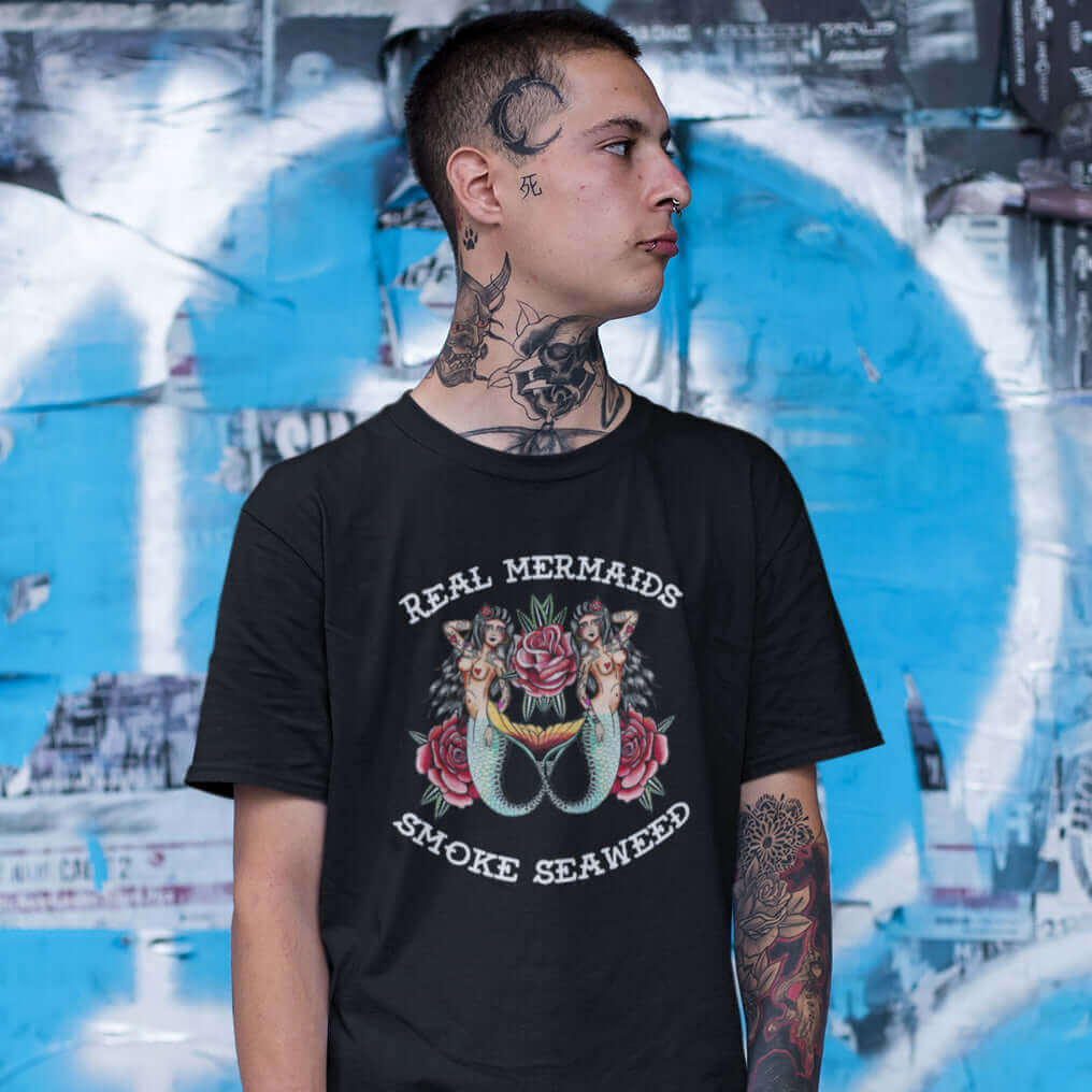 Tattooed man wearing a black t-shirt with image of 2 mermaids and the words Real mermaids smoke seaweed printed on the front.