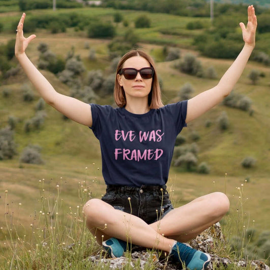 Woman sitting on a hill wearing navy blue t-shirt with the words Eve was framed printed on the front in pink.