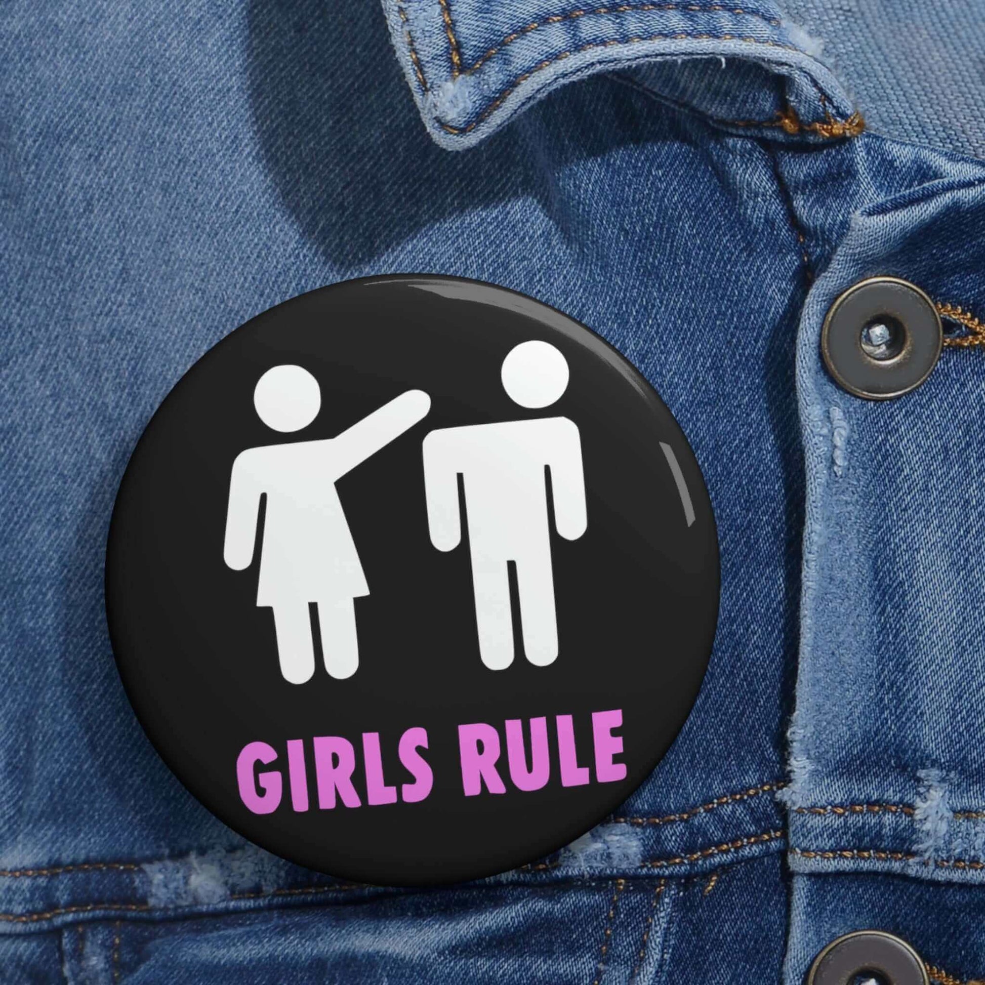 Black pin-back button with white stick figure man and woman. The woman is punching the mans head off and the words Girls rule is printed beneath in hot pink.