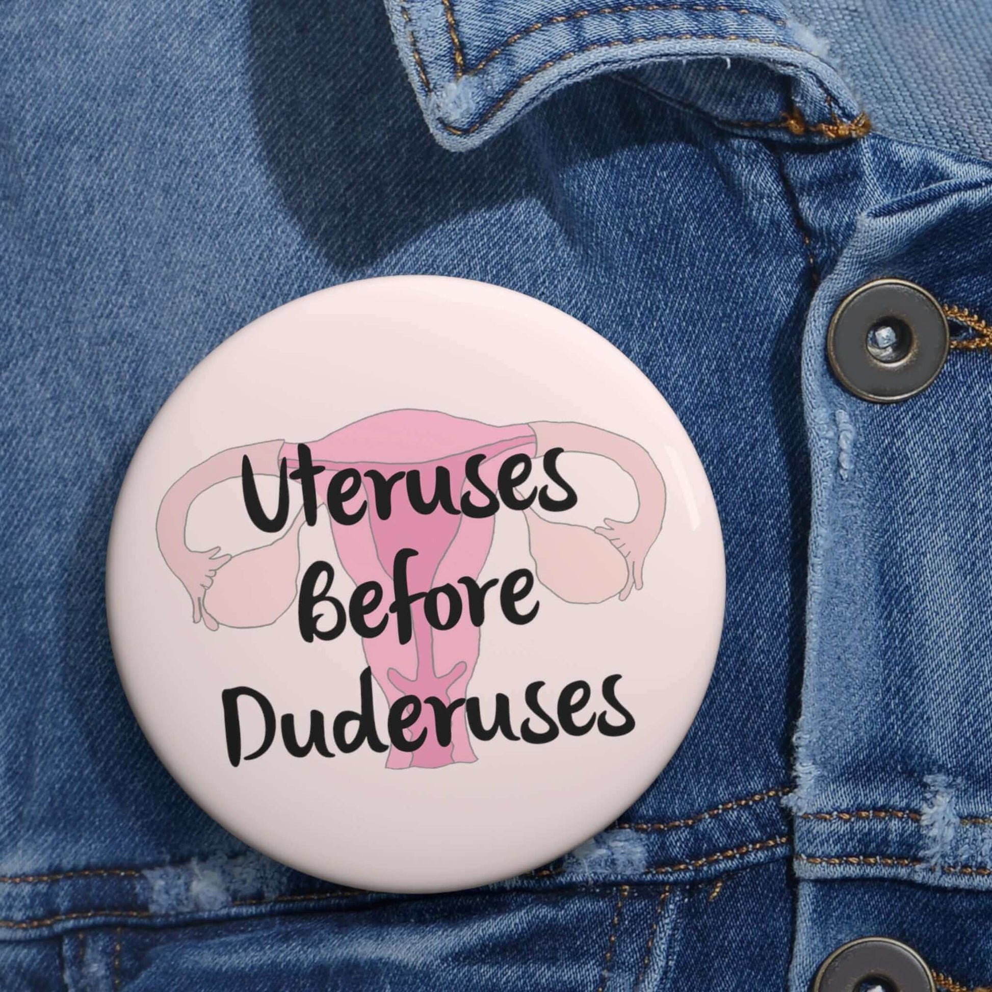 Pink pinback button with image of a uterus and the words Uteruses before duderuses.