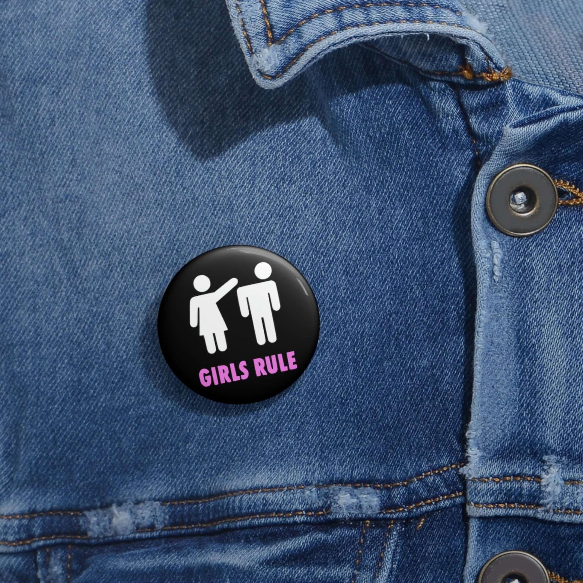 Black pin-back button with white stick figure man and woman. The woman is punching the mans head off and the words Girls rule is printed beneath in hot pink.