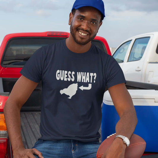 Man sitting on the tailgate of a truck. He is wearing a navy blue t-shirt with an image of a chicken and the words Guess what question mark. There is an arrow pointing to the chickens butt. The graphics are printed on the front of the shirt.