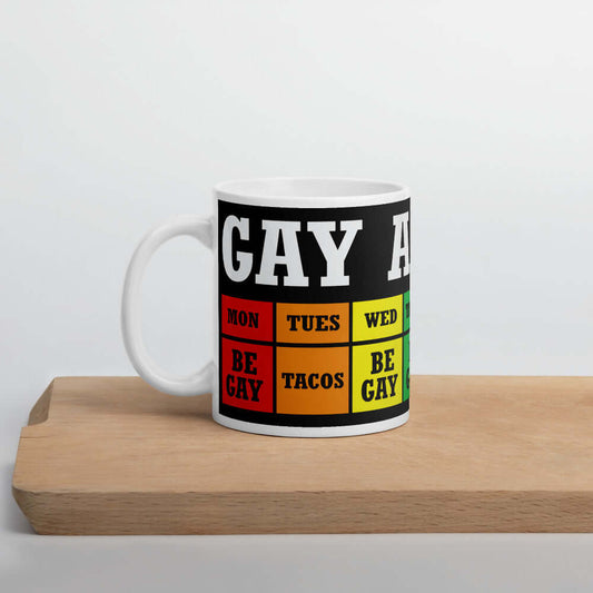 White ceramic coffee mug with funny Gay Agenda weekly calendar printed around the mug. The week is all in different rainbow colors. Monday says Be gay. Tuesday says Tacos. Wednesday & Thursday say Be gay. Friday & Saturday say Super gay and Sunday says br