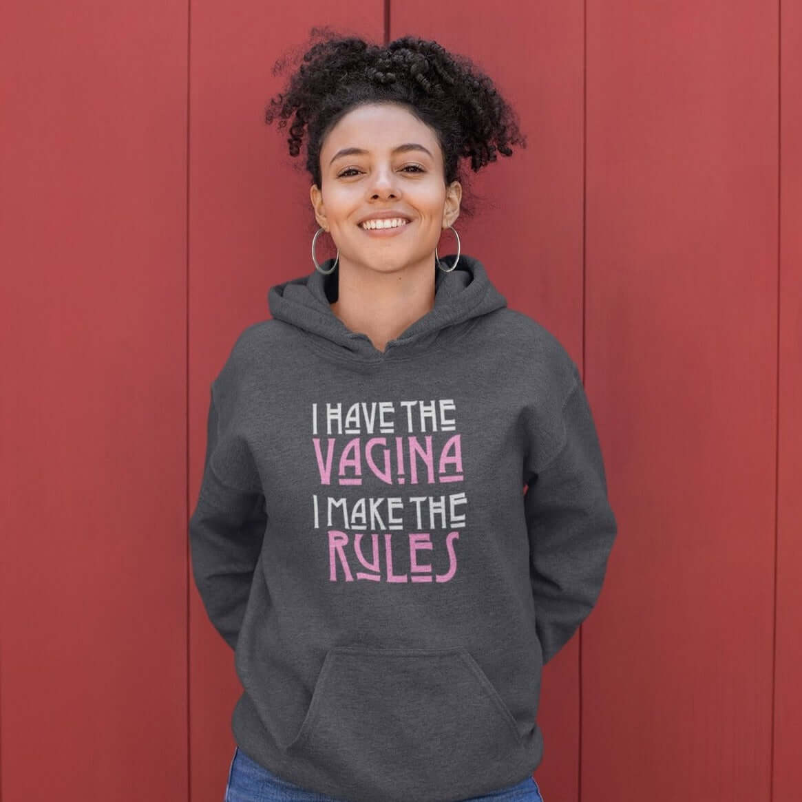 Woman wearing dark heather grey hoodie sweatshirt with the words I have the vagina , I make the rules printed on the front. The words vagina and rules are pink, the rest of the words are white.