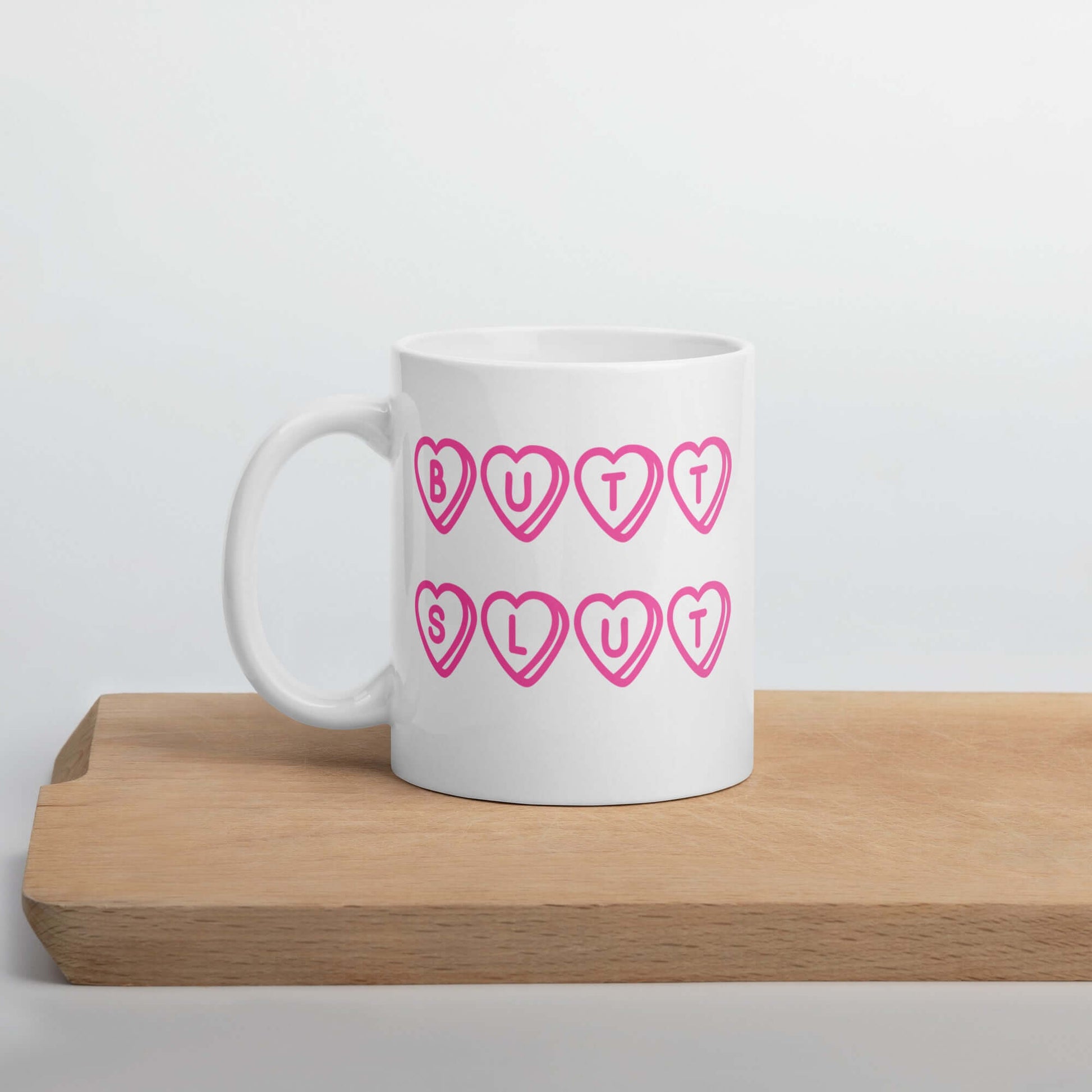 White ceramic mug with the words Butt Slut printed in pink on both sides. Pink hearts are around each letter in the text.