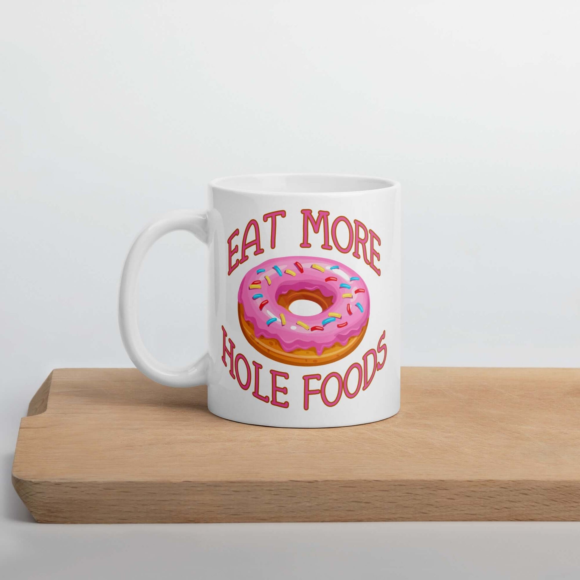 White coffee mug with an image of a donut with pink icing and sprinkles and the words Eat more hole foods printed on both sides.