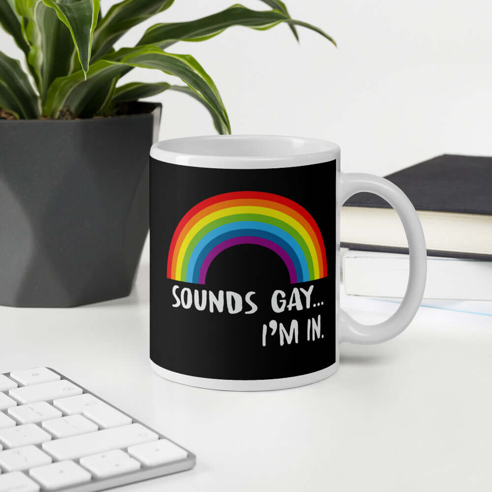 Black and white ceramic coffee mug that has an image of a rainbow and the phrase Sounds gay, I'm in printed on both sides.