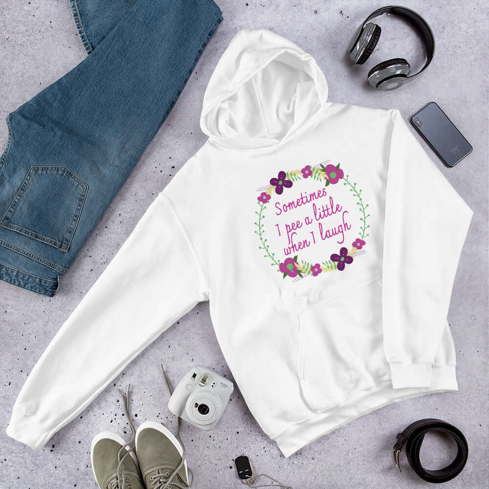 White hoodie sweatshirt that has a minimalistic style line drawing floral wreath with the phrase Sometimes I pee a little when I laugh printed in the center of the wreath. The graphics are printed on the front of the hoodie.