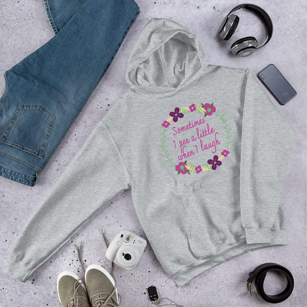 Light grey hoodie sweatshirt that has a minimalistic style line drawing floral wreath with the phrase Sometimes I pee a little when I laugh printed in the center of the wreath. The graphics are printed on the front of the hoodie.