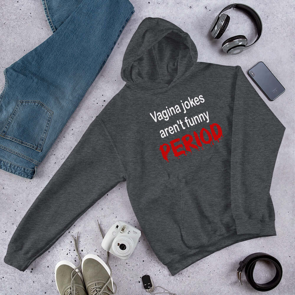 Dark heather hoodie sweatshirt with the crude phrase Vagina jokes aren't funny...period. The word period is in a red drippy font. The graphics are printed on the front of the hoodie.