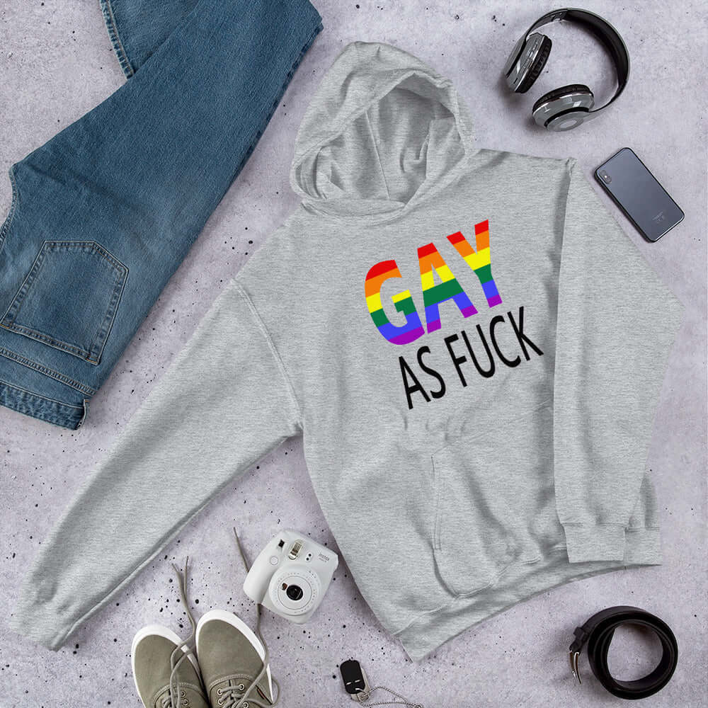 Light grey hoodie sweatshirt with the words Gay as fuck printed on the front. The word Gay is in rainbow stripe font.