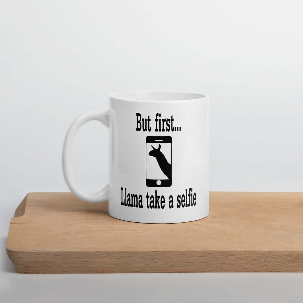 White ceramic coffee mug with image of an outline of a cell phone with image of a llama on the screen. The words But first...llama take a selfie are around the image. The graphic is printed on both sides of the mug,