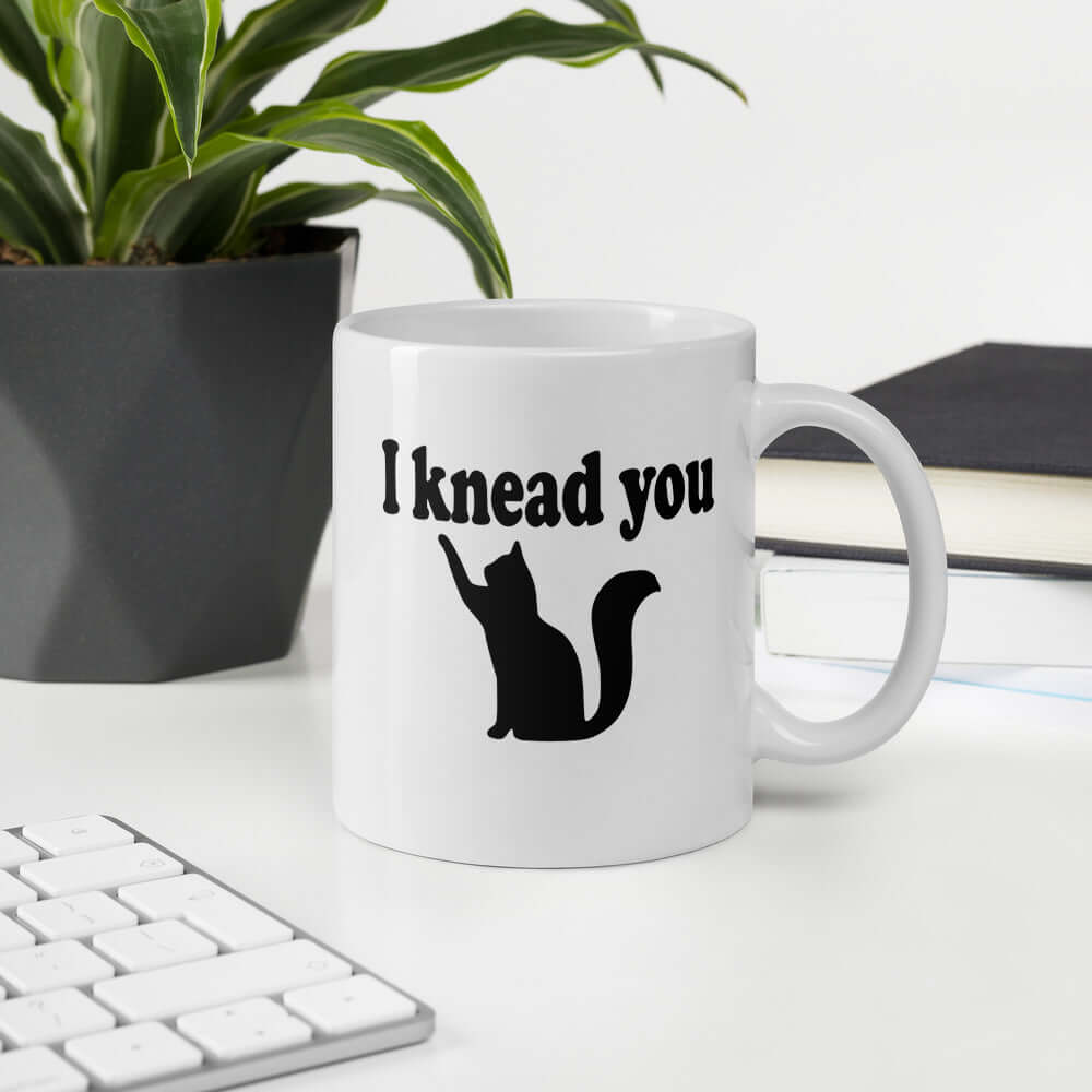 White ceramic coffee mug that has a pun image of a silhouette of a cat and the words I knead you printed on both sides of the mug.
