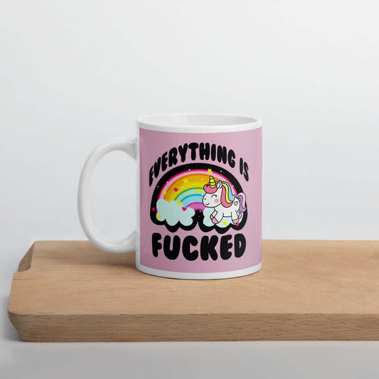 White ceramic coffee mug with a graphic of a kawaii style unicorn and a pastel rainbow with the words Everything is fucked printed on both sides. The background is pink.