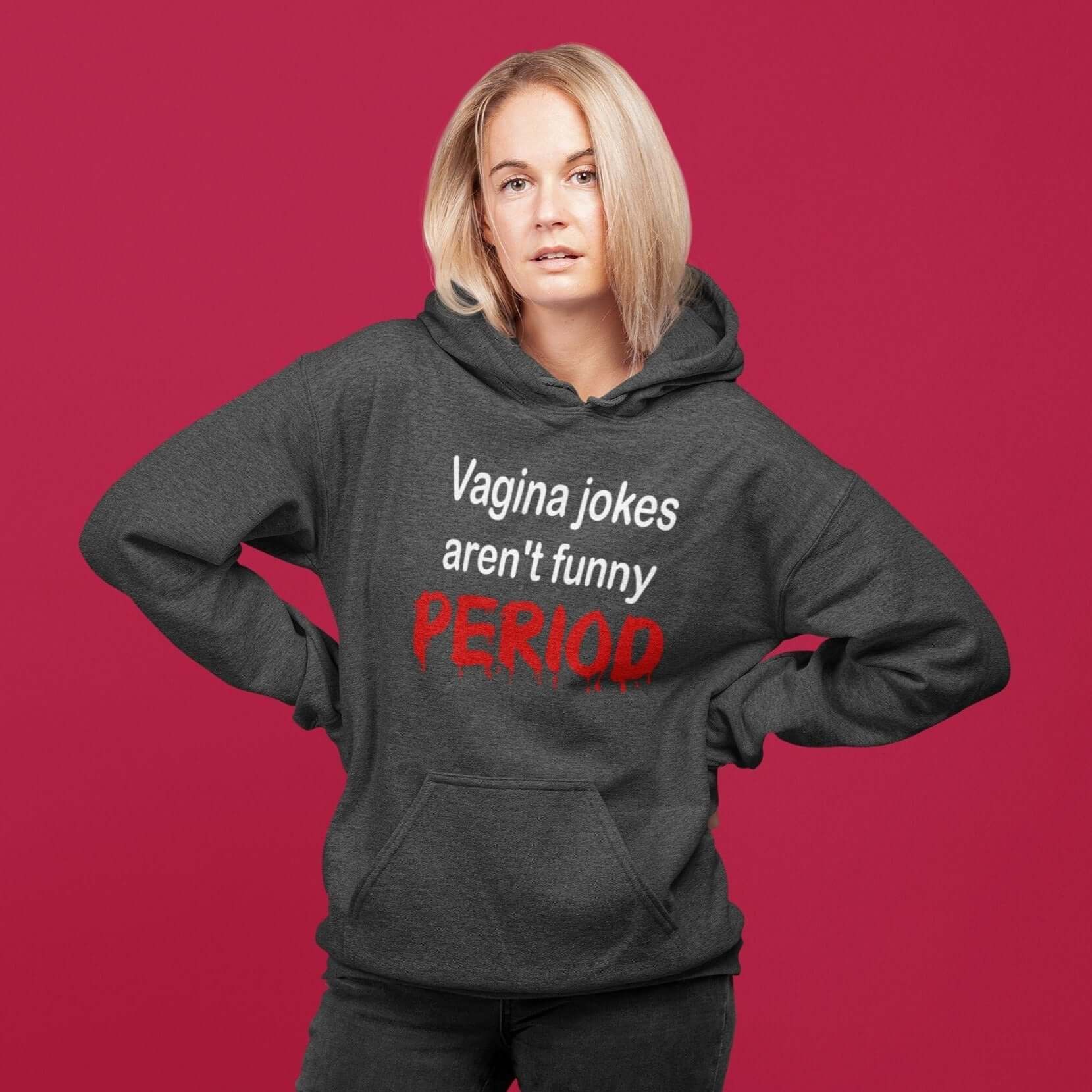 Sassy looking woman wearing a dark heather grey hoodie sweatshirt with the crude phrase Vagina jokes aren't funny...period. The word period is in a red drippy font. The graphics are printed on the front of the hoodie.