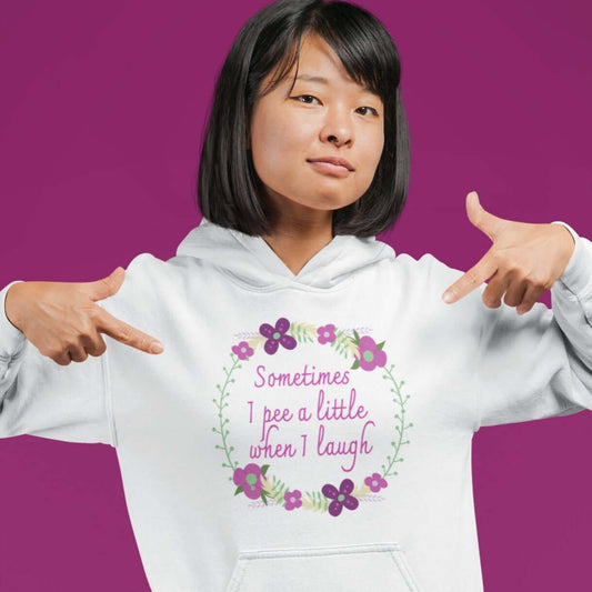 Woman wearing a light pink hoodie sweatshirt. The hoodie sweatshirt has a minimalistic style line drawing floral wreath with the phrase Sometimes I pee a little when I laugh printed in the center of the wreath. The graphics are printed on the front of the