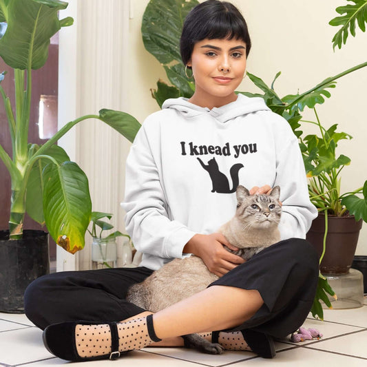 Woman petting a cat while wearing a white hoodie sweatshirt that has a pun image of a silhouette of a cat and the words I knead you printed on the front.