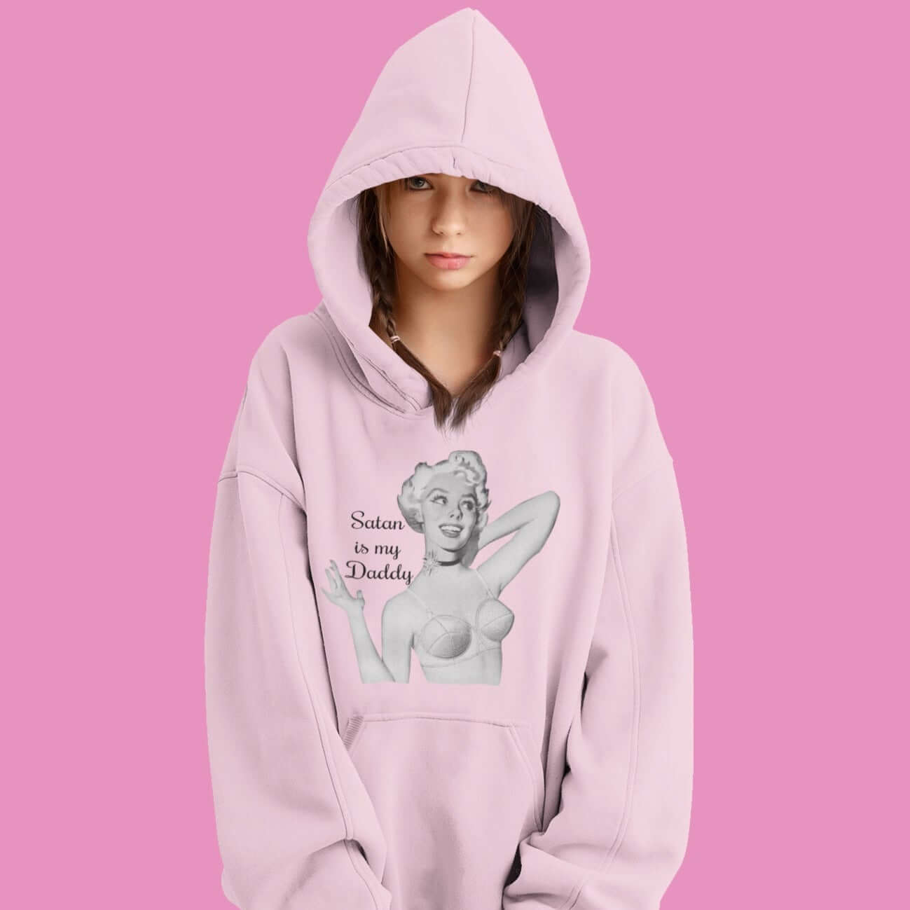 Woman wearing a light pink hoodie sweatshirt with the hood up and partially covering her face. The hoodie has an image of a retro black & white pin-up model and the phrase Satan is my Daddy printed on the front.