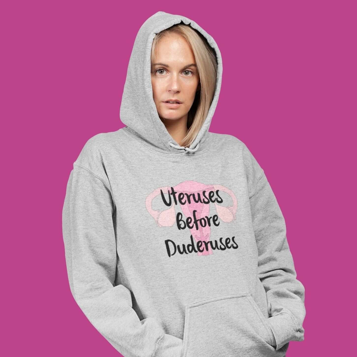 Woman wearing a light grey hoodie sweatshirt with image of a uterus and the words Uteruses before duderuses printed on the front.