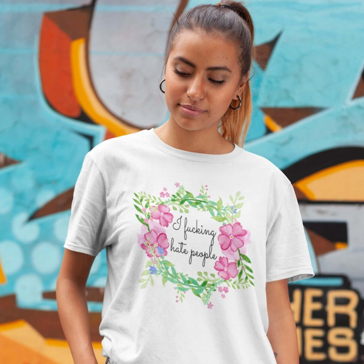 Woman wearing white t-shirt with pink and green floral wreath image and the words I fucking hate people printed on the front.