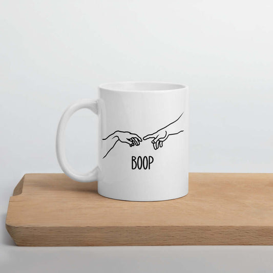 White ceramic mug with line drawing hands reaching out and touching in the center like they in the famous art creation of Adam. The word Boop is printed underneath.