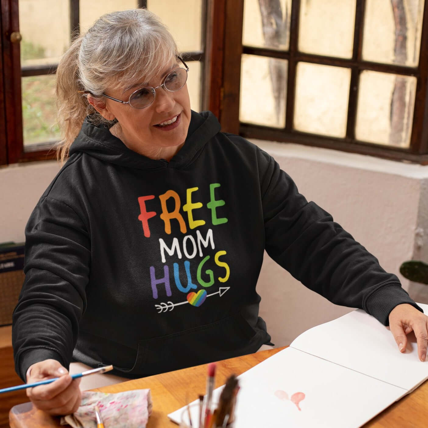 Older woman wearing black hooded sweatshirt with the words Free Mom hugs in rainbow lettering printed on the front.