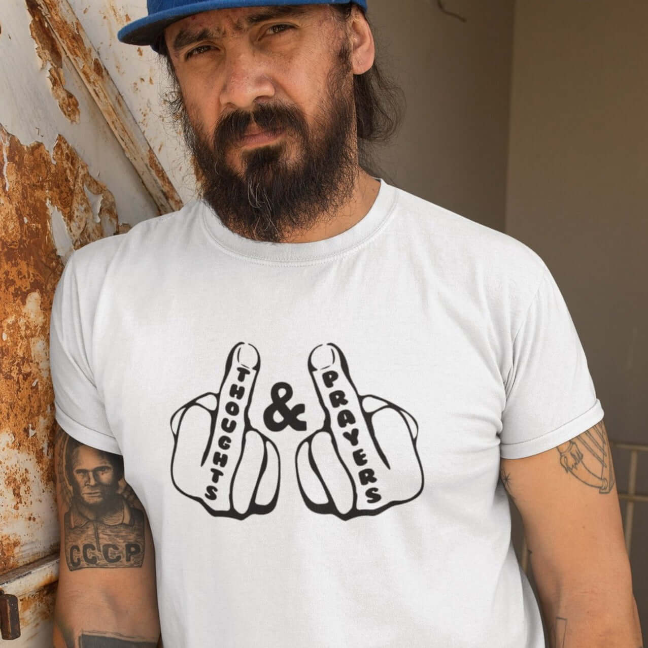 Man leaning against a wall wearing a white t-shirt. The t-shirt has an image of 2 line drawing hands with the middle fingers up. The word Thoughts is printed on one middle finger and the word Prayers is printed on the other middle finger. The graphics are