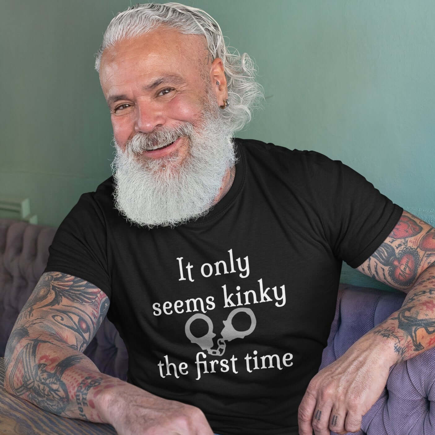 Bearded older man wearing a black t-shirt with the phrase It only seems kinky the first time printed on the front. There is an image of handcuffs with the text.