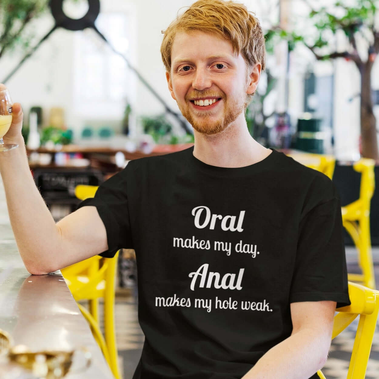 Man sitting at a cafe table wearing a black t-shirt with the pun phrase Oral makes my day Anal makes my hole weak printed on the front.