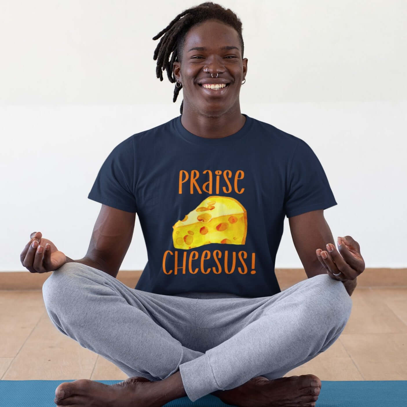 Smiling man meditating wearing a navy blue t-shirt with funny graphics of a piece of swiss cheese and the words Praise Cheesus printed on the front of the hoodie in yellow and orange.