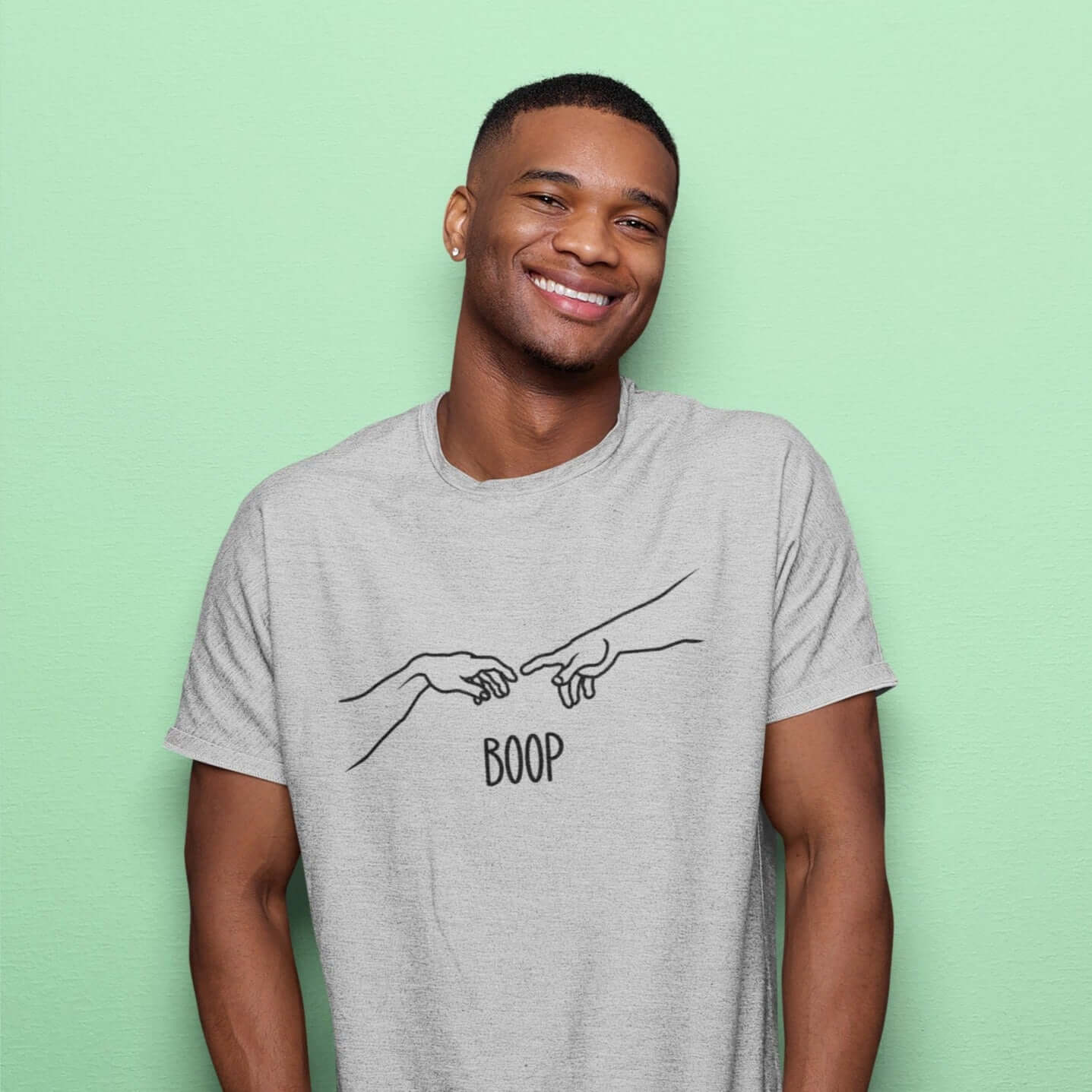 Smiling man wearing a light grey t-shirt with line drawing hands reaching out and touching in the center like they in the famous art creation of Adam. The word Boop is printed underneath.