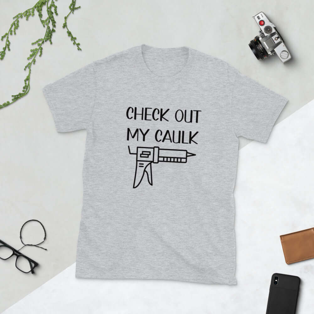 Light grey t-shirt with the pun phrase Check out my caulk with a line drawing image of a caulking gun printed on the front.