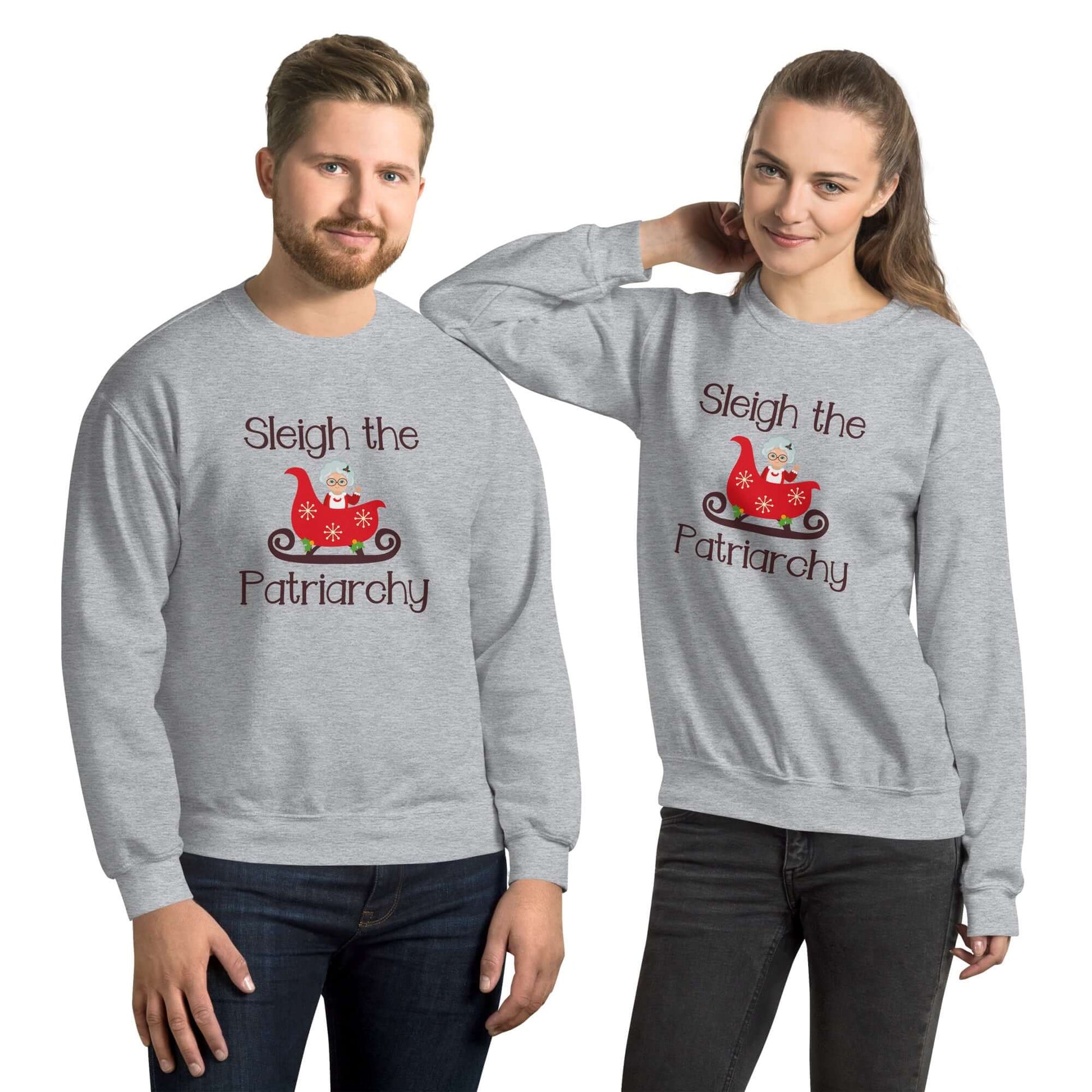 Man and a woman standing next to each other. Both are wearing a light grey crewneck sweatshirt with graphic of Mrs. Claus sitting in a sleigh and the words Sleigh the patriarchy printed on the front.