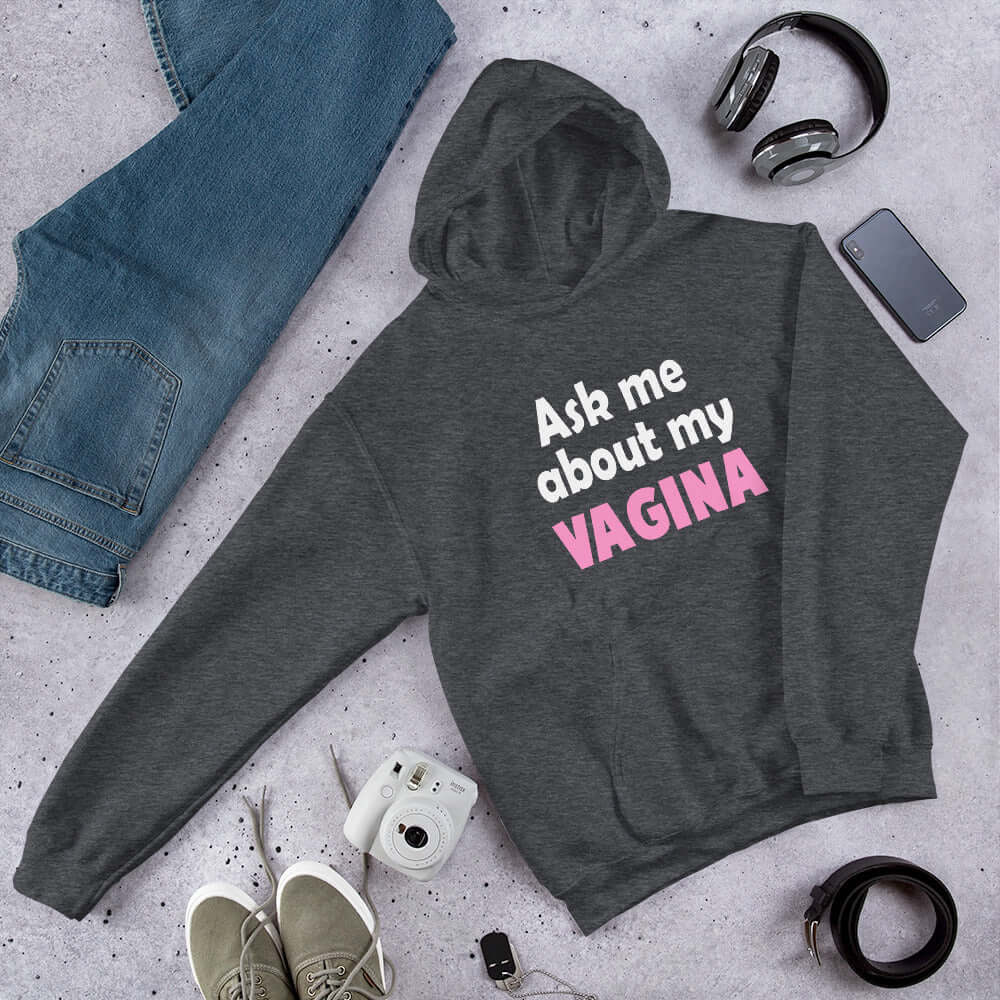 Dark heather grey hoodie sweatshirt with the words Ask me about my vagina printed on the front. The word vagina is printed in pink. The rest of the text is white.