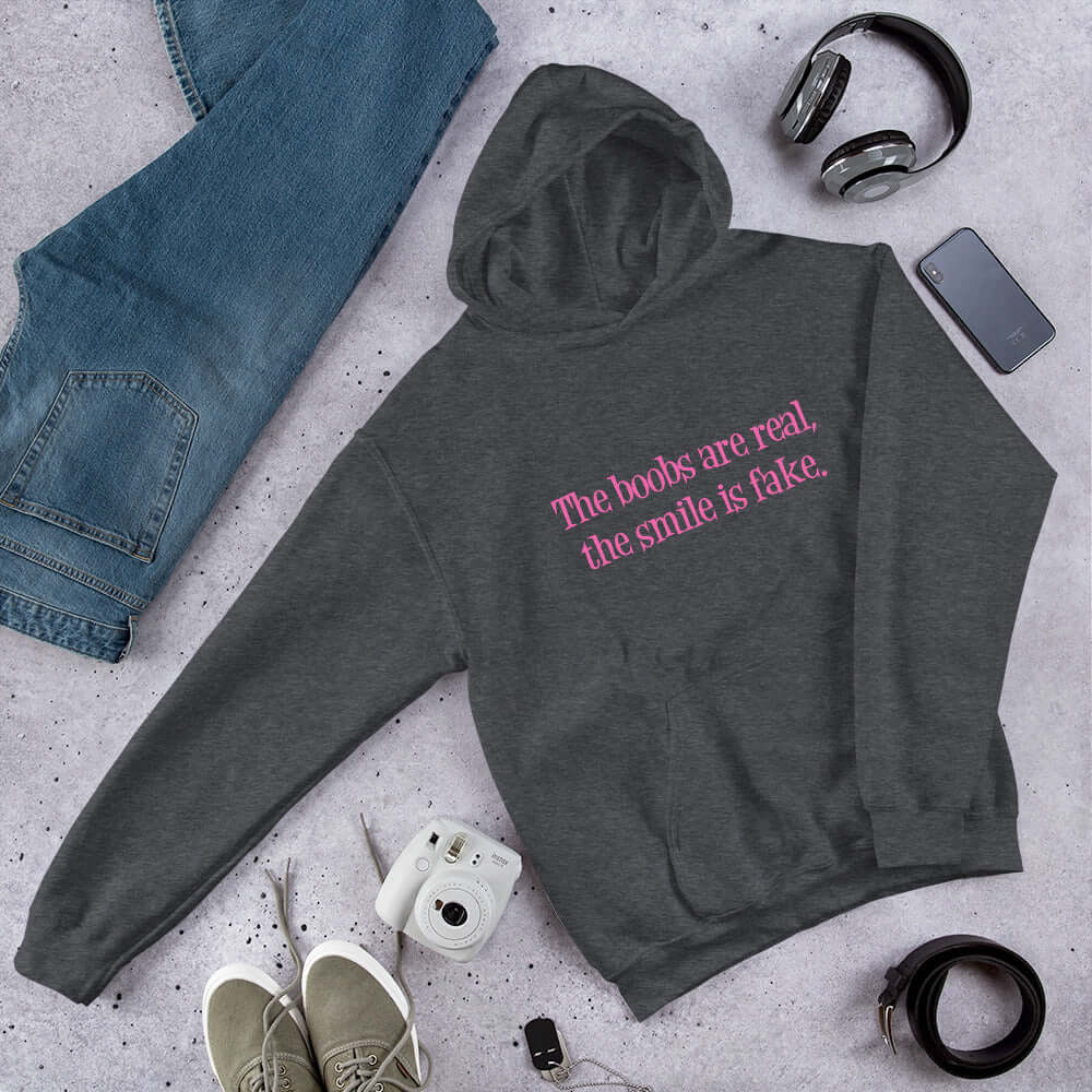 Dark heather grey hoodie sweatshirt with the phrase The boobs are real, the smile is fake printed in pink on the front.