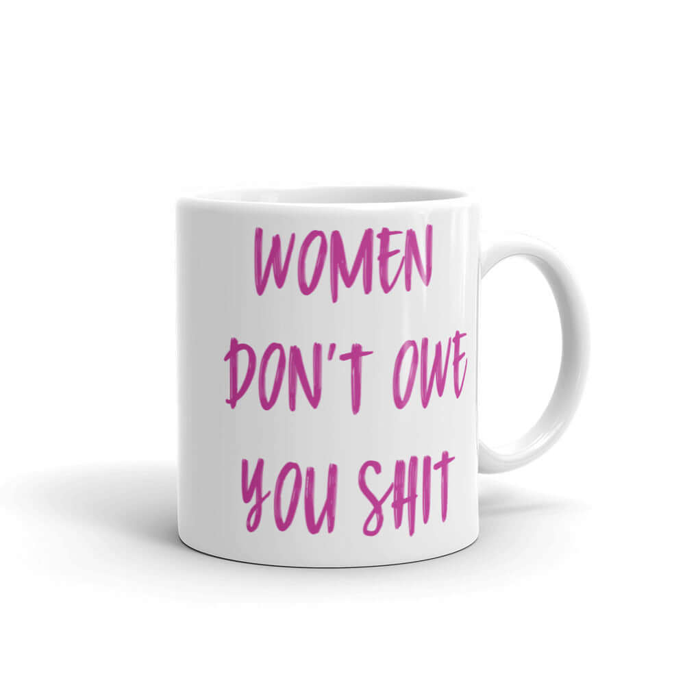 White ceramic coffee mug with the words Women don't owe you shit printed on both sides in pink.