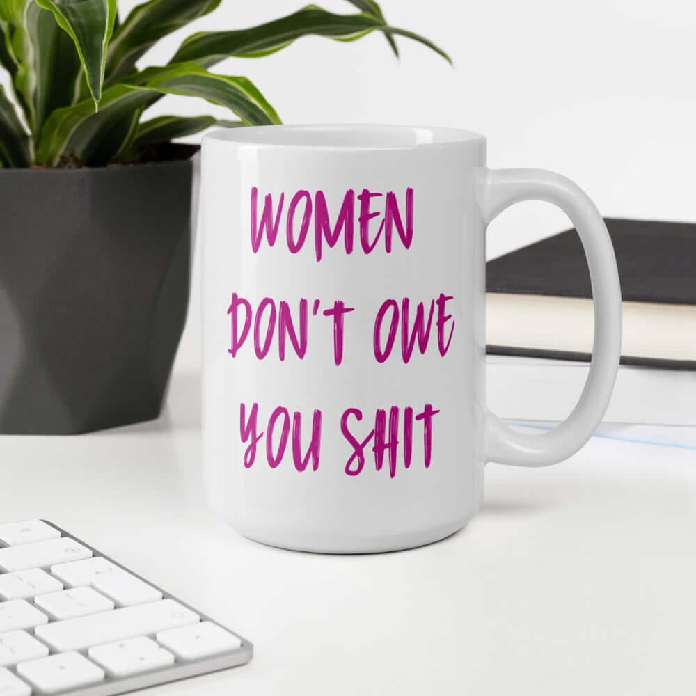 White ceramic coffee mug with the words Women don't owe you shit printed on both sides in pink.