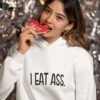 Woman biting a red cookie wearing a white hoodie sweatshirt with the text 'I eat ass' printed on the front.
