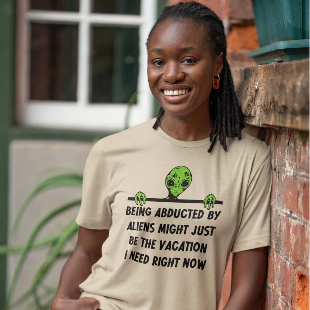 Woman wearing a sand color alien abduction joke t-shirt with image of an alien and the words 'Being abducted by aliens might just be the vacation I need right now' printed on the front.