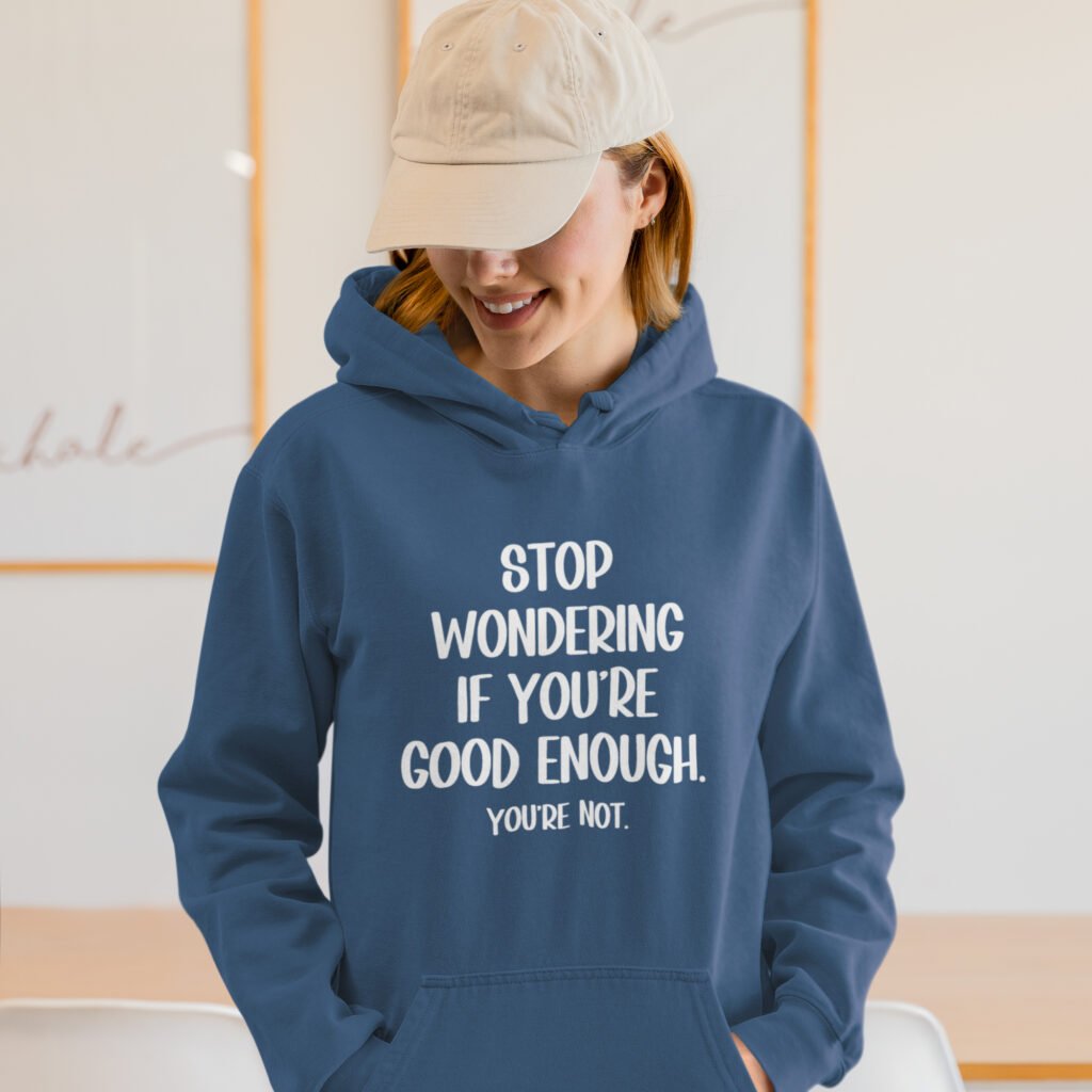 Woman wearing an indigo blue hoodie sweatshirt with the phrase 'Stop wondering if you're good enough. You're not.' printed on the front. The words 'You're not' are smaller then the rest.