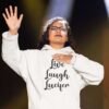 Woman praising while wearing a white hoodie sweatshirt with the parody phrase 'Live, Laugh, Lucifer' printed on the front.