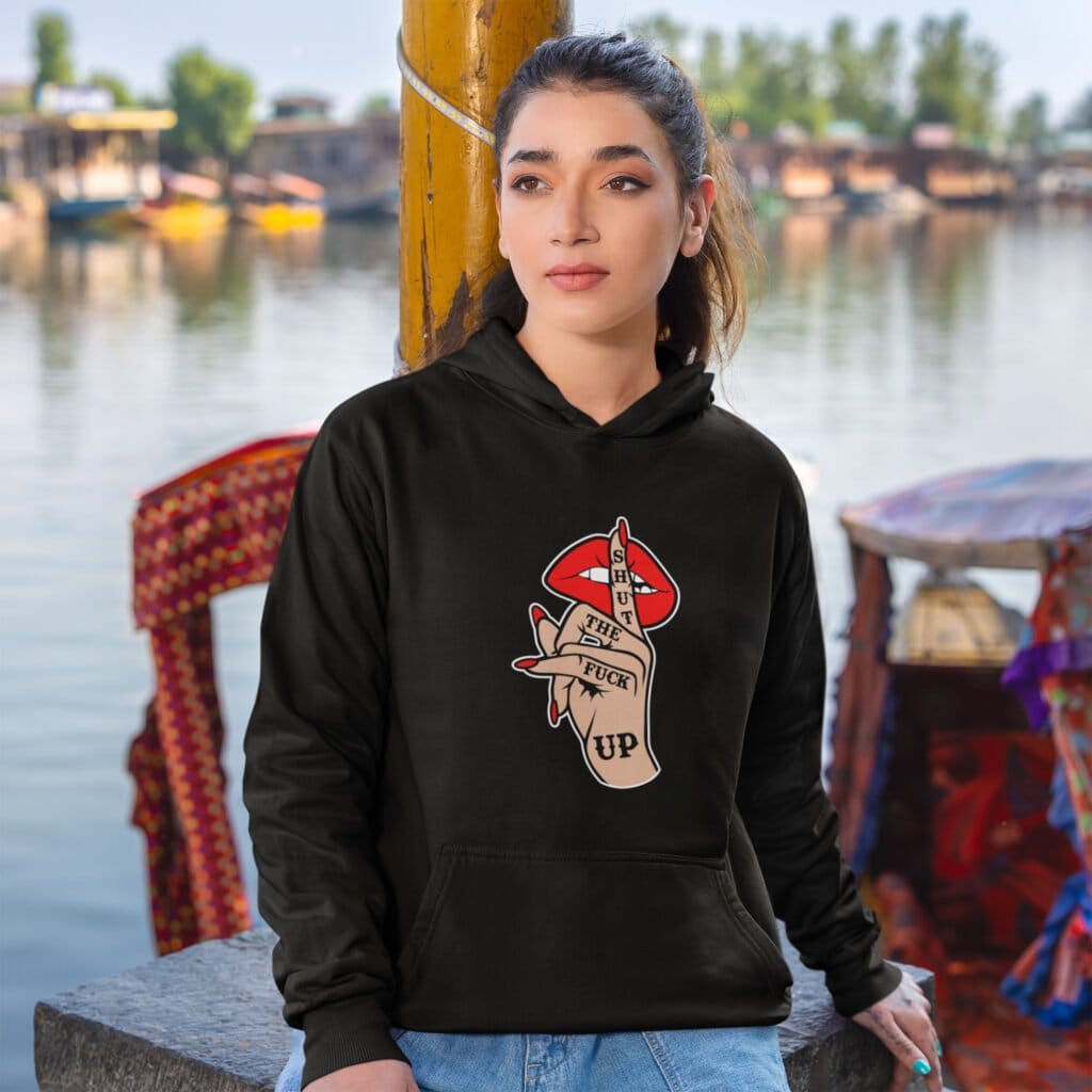 Woman wearing a black hoodie sweatshirt with an image of a hand over lips making the shh gesture. The words 'Shut the fuck up' are printed on the hand. The graphics are printed on the front.