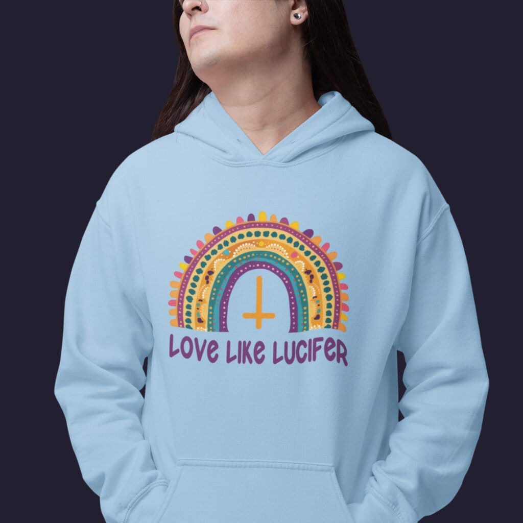 Man wearing a light blue hoodie sweatshirt with an image of a boho style rainbow. There is an upside down cross in the center arch of the rainbow and the text 'Love like Lucifer' printed under. The graphics are printed on the front.